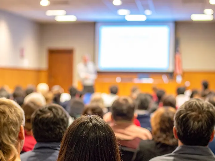Audience seated from behind watching a presenter give a slide presentation at a town hall meeting